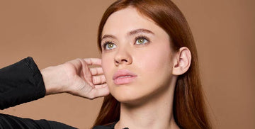 Close-up of a young red-haired woman wearing a black long-sleeve shirt, touching her face with her right arm.