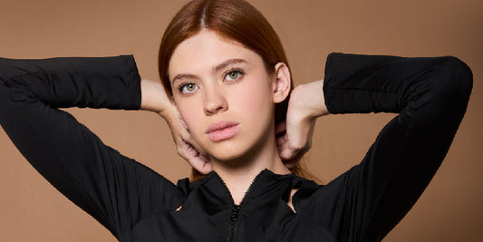 Close-up of a young red-haired woman wearing a black long-sleeve shirt, with her arms up resting behind her head.