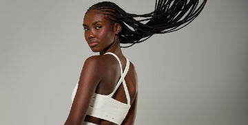 A young African-American woman wearing a white sports bra, looking at the camera and flipping her hair, on a grey background.