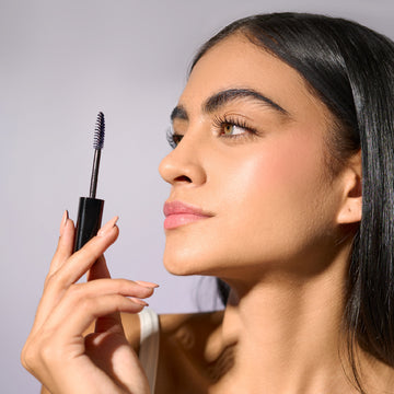 Close-up of a young woman's face with dark hair, holding the mascara brush of Motion Beauty Sport Lash Waterproof Mascara.