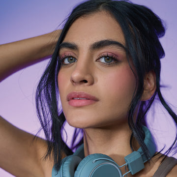 Close-up of a young woman with headphones around her neck, holding her black hair up, looking at the camera.